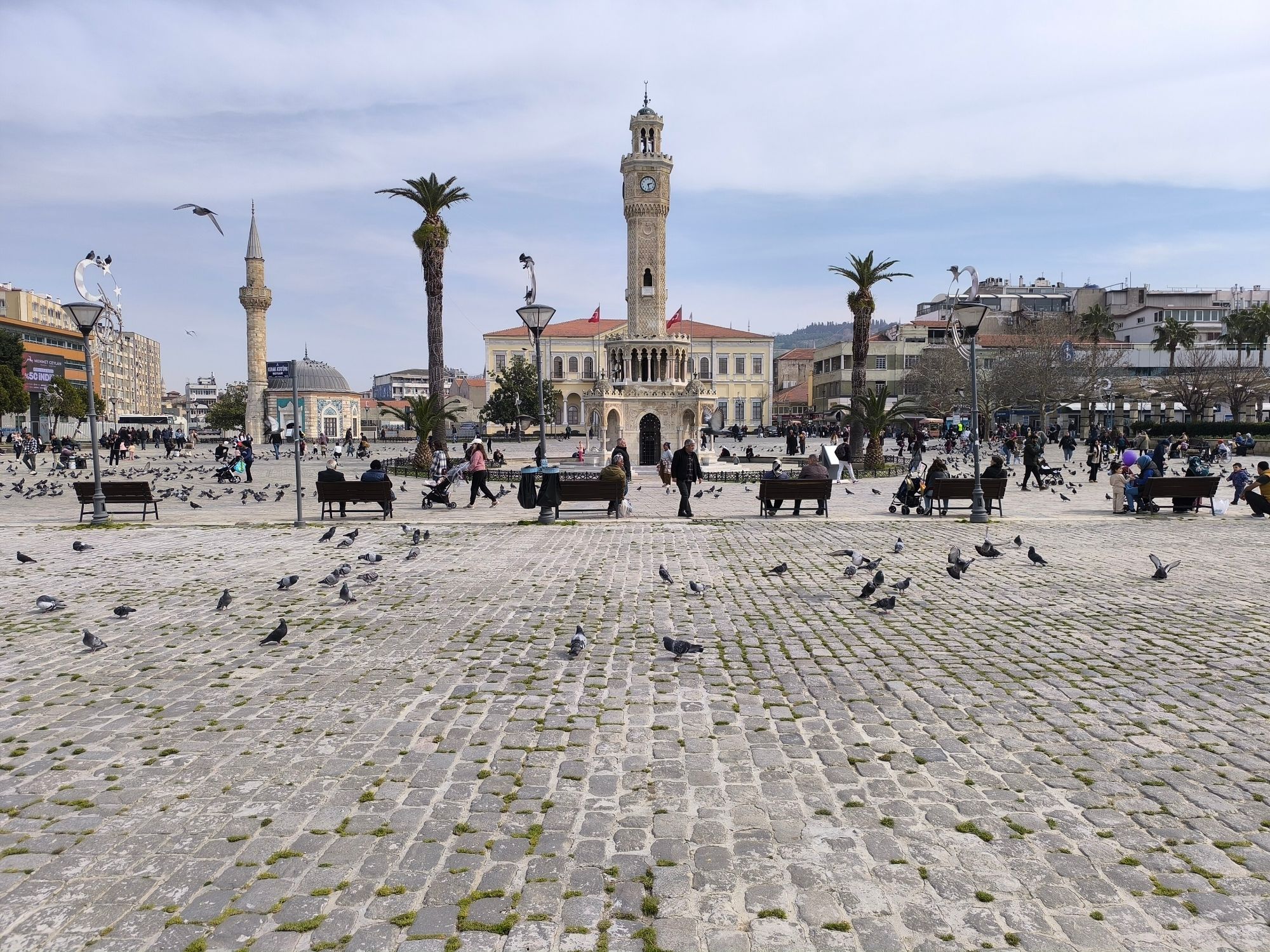 Konak Square in İzmir with its Ottoman clock tower, palm trees and pigeons wheeling over the paving